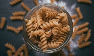 a scoop of brown corkscrew-like pasta in a jar held over more pasta and flour on the counter below